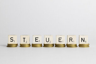 Das Wort "Steuern" geschrieben mit Scrabble-Steinen. Jeder Stein steht auf einem Stapel von 3 Geldstücken.